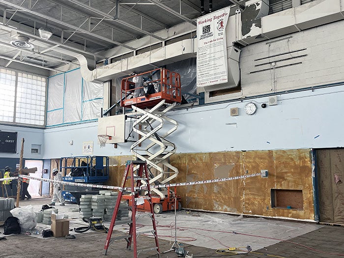 Interior of a gymnasium undergoing renovation, with workers on a scissor lift repairing overhead ductwork. The gym walls are partially stripped, protective plastic sheeting covers the windows, and construction materials and tools are scattered around the floor beneath a basketball hoop.