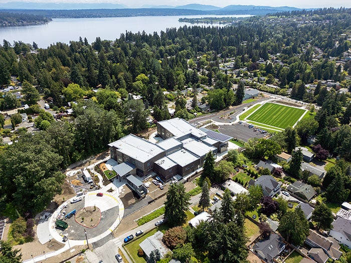 An aerial photograph of a large, modern school campus surrounded by trees and residential neighborhoods, with a scenic lake and distant mountains in the background. The school buildings are centered in the image, and a natural turf sports field is visible on the right.