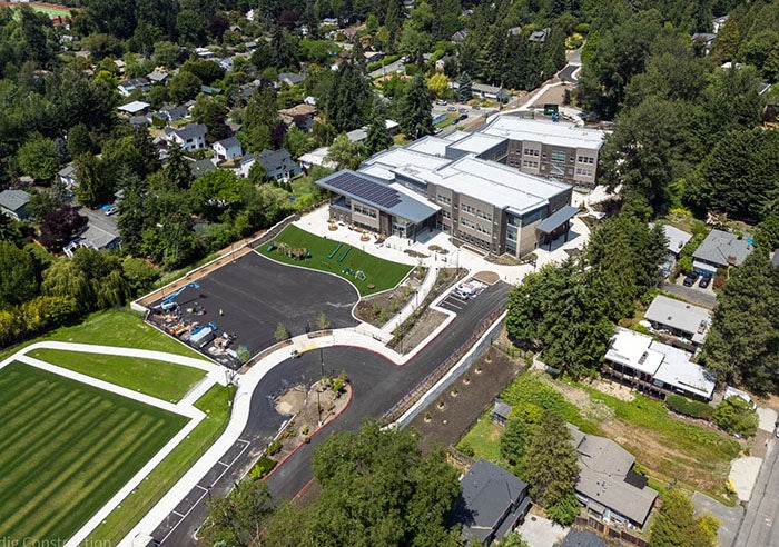 A top-down view of a newly constructed school with angular building wings, solar panels on the roof, and landscaped grounds. A blacktop parking area and green athletic field are included, with surrounding trees and homes lining the perimeter.