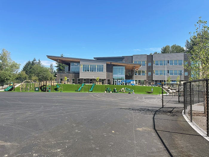 Ground-level view of a three-story school building with a sweeping roofline and a fenced playground in front. The play area features green equipment and turf hills, set against a backdrop of clear blue sky and trees.