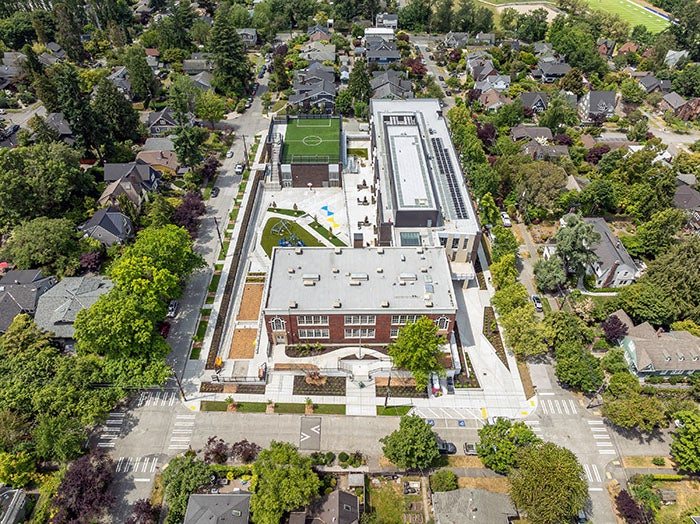 Aerial view of a modernized elementary school campus surrounded by residential streets and trees. The school buildings are arranged in a long rectangular layout, featuring a playfield with a soccer court, outdoor play areas, and landscaped walkways.