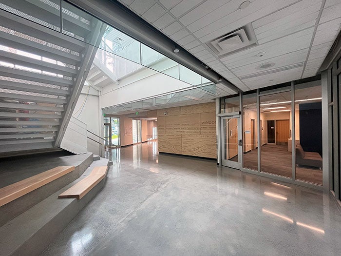 Interior of a school entry lobby with polished concrete floors, a modern staircase with wood seating steps, glass walls, and a welcoming mural on the back wall. Natural light streams in through skylights and tall windows.