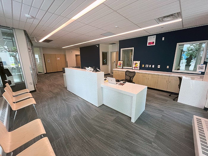 A clean, modern school reception area with a white front desk, blue accent wall, office chairs, and large windows. The space includes visitor seating with wooden chairs and a carpeted floor with gray tones.
