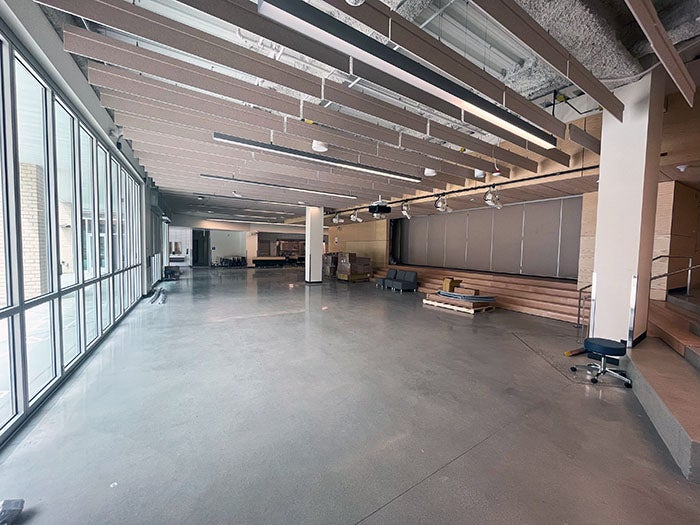 A spacious multipurpose commons area with floor-to-ceiling windows along one side, overhead acoustic ceiling baffles, built-in stage seating, and polished concrete flooring. Furniture and materials are arranged throughout the open space.