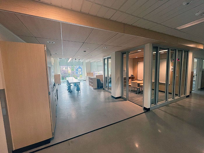 An open classroom breakout space with glass-walled rooms, a central group table, shelving, and natural light coming through large windows. Light wood tones and soft lighting create a welcoming environment.