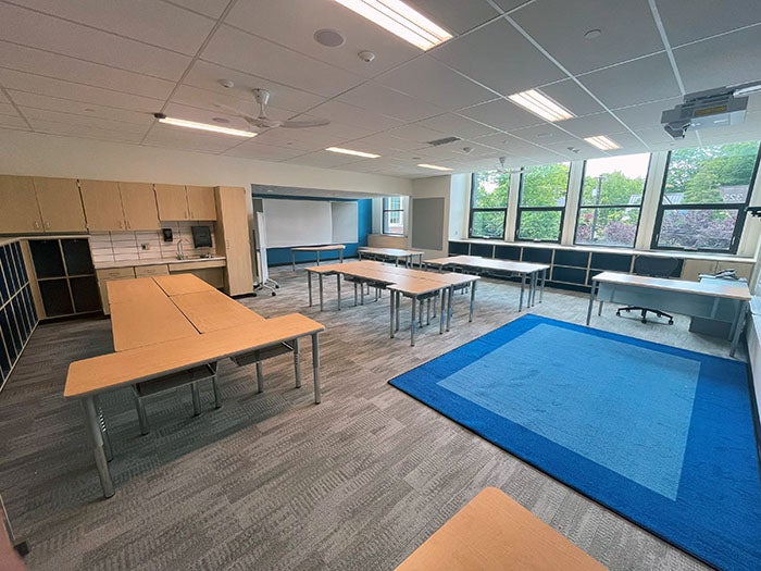 A large, modern classroom with individual and grouped tables, built-in storage cabinets, a blue rug, and large windows that provide ample daylight. The room includes whiteboards and technology for instruction.