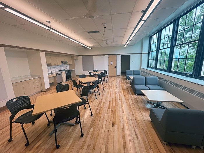 A staff lounge with light wood flooring, soft seating, dining tables, a kitchenette with appliances, and large windows providing outdoor views. The space is well-lit and designed for comfort and collaboration.