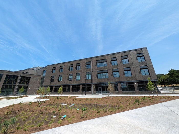 Wide-angle view of a three-story school building with a dark brick exterior and large windows. The foreground includes a landscaped area with young trees and newly planted greenery under a clear blue sky.
