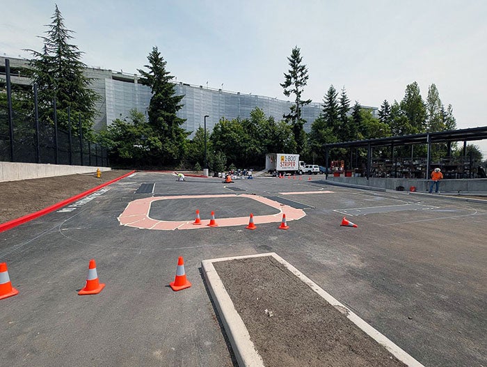 Freshly paved play area marked with painted shapes and surrounded by orange construction cones. Workers are visible in the distance preparing the surface. A large, multi-story building is in the background behind a row of trees.