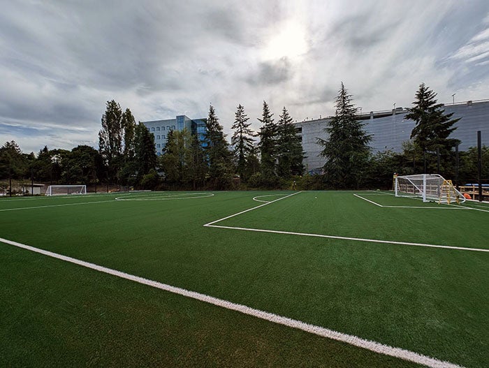 A large, bright green artificial turf soccer field with white markings and multiple goal posts. The area is enclosed by tall trees and modern office buildings, with a cloudy sky overhead.