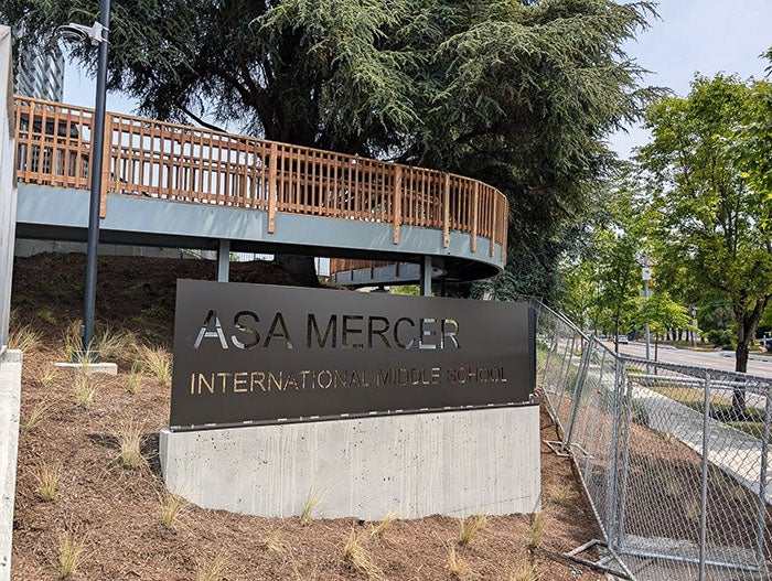 Sign for “Asa Mercer International Middle School” mounted on a concrete base at the edge of a fenced campus. Behind the sign is a raised, curved walkway with a wooden railing, and large trees providing shade.