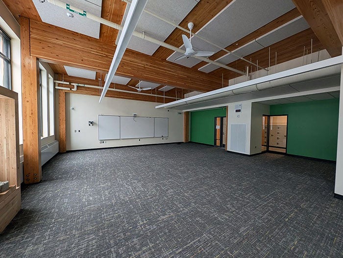 Spacious, carpeted classroom with natural wood ceiling beams, acoustic panels, and overhead lighting. The walls feature whiteboards and green accent paint, and a door opens to an adjacent hallway with lockers.