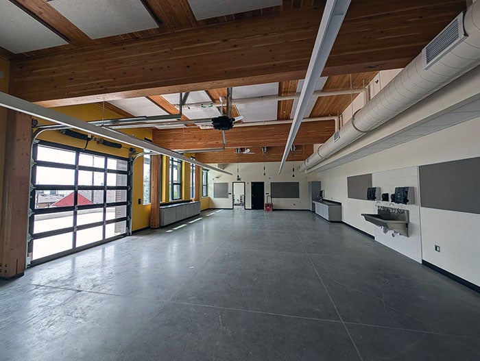 Modern learning space with a polished concrete floor, exposed wood ceiling, and a large roll-up glass garage door. The room includes workstations, wall-mounted sinks, and natural light from tall windows.