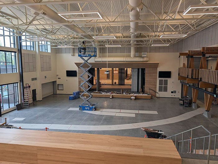 Large multipurpose space with high ceilings, a built-in stage, and a scissor lift where a worker is adjusting ceiling fixtures. The room includes seating platforms.