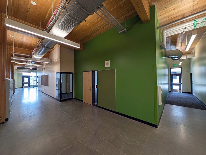 Interior corridor with polished concrete floors, exposed ceiling ducts, and a bright green wall with classroom doors. Natural light enters from both ends of the hallway, and utility piping is visible overhead.