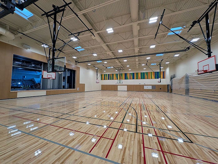 Spacious school gymnasium with a glossy hardwood basketball court, retractable hoops, and ceiling-mounted lights. The back wall features colorful acoustic panels, and a stage with curtains is visible to the left.