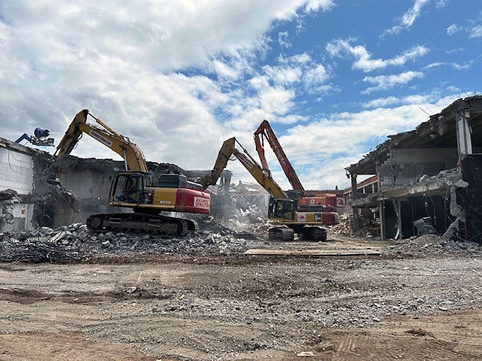 Three large excavators are actively demolishing a multi-story building, surrounded by piles of concrete rubble and debris. Dust fills the air as the structure collapses, and the sky above is partly cloudy with blue patches showing through.