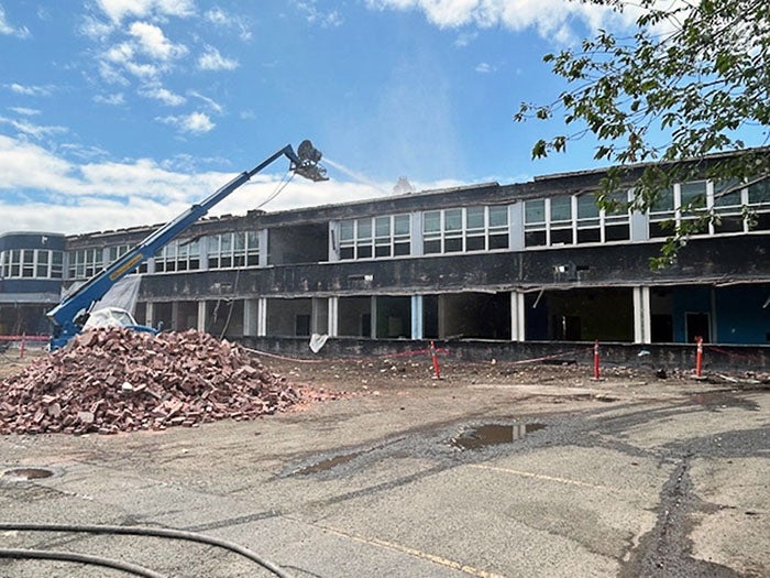 A partially demolished two-story school building with shattered windows and an exposed interior. A high-reach water spray rig is actively reducing dust while a large pile of red bricks sits in the foreground on the cracked pavement.