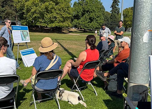 outdoor meeting with one person standing with presentation board on easel and a group of people facing them in chairs