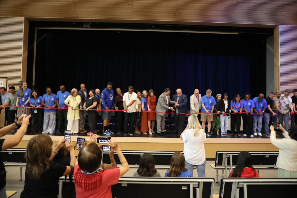 a group of people are lined up across a stage with a red ribbon in front of them. the ribbon has just been cut and hasn't fallen yet