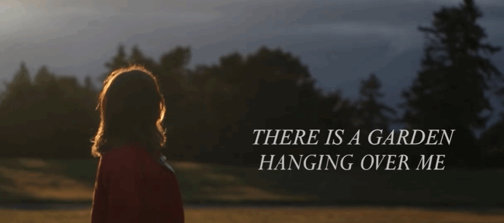 still from film of person walking with text THERE IS A GARDEN HANGING OVER ME