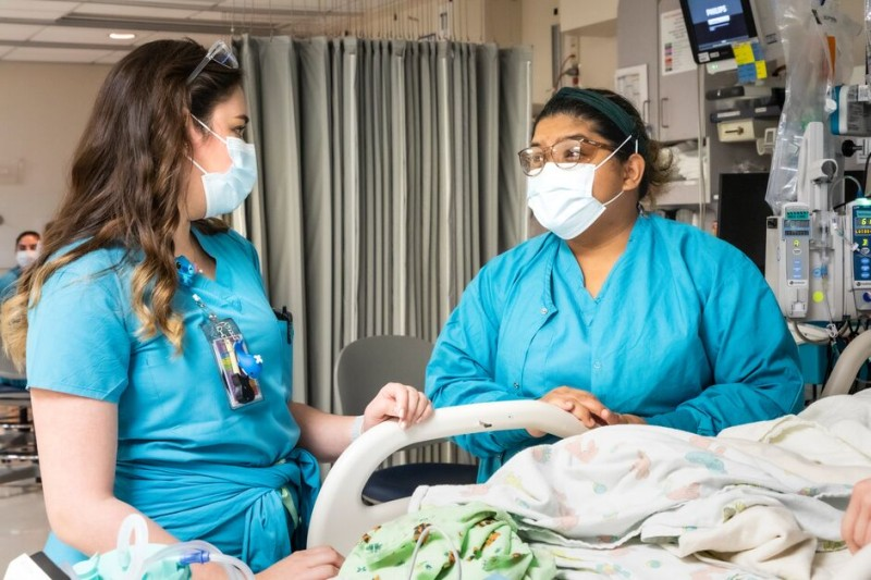A nurse working with a summer camp student, both in blue scrubs, in a hospital room