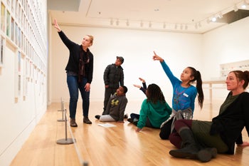 Students point to an art work with a teacher in a gallery