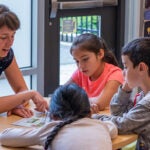 A teacher talks with three young students at a table.