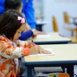 A young student sits at a desk with a pencil in her hand.