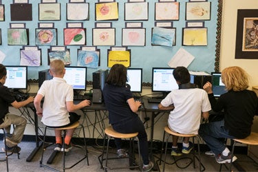 Five students work at computers side by side in a classroom