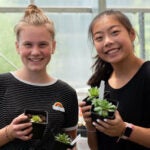 Two high school students smile for a photo while holding small plants in a greenhouse.