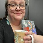 A woman whose hands are full with a laptop and a coffee mug makes a friendly smirk to the camera. It looks like she's in an elevator.