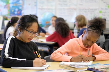 Two students work side by side at a desk in a classroom