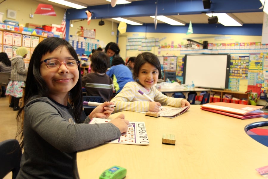 Two students smile for a photo while sitting at a desk in a classroom