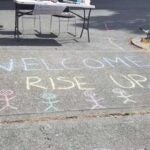 A sidewalk on school grounds with a table and kid's bike. Sidewalk chalk written 