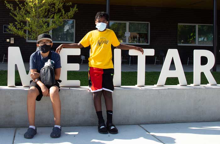 Two students stand together outside wearing masks in front of a school