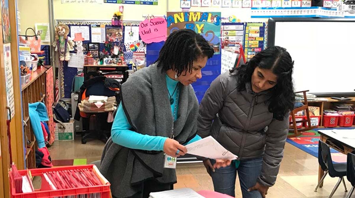 A teacher and adult stand in a classroom discussing a paper.
