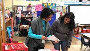 A teacher and adult stand in a classroom discussing a paper.