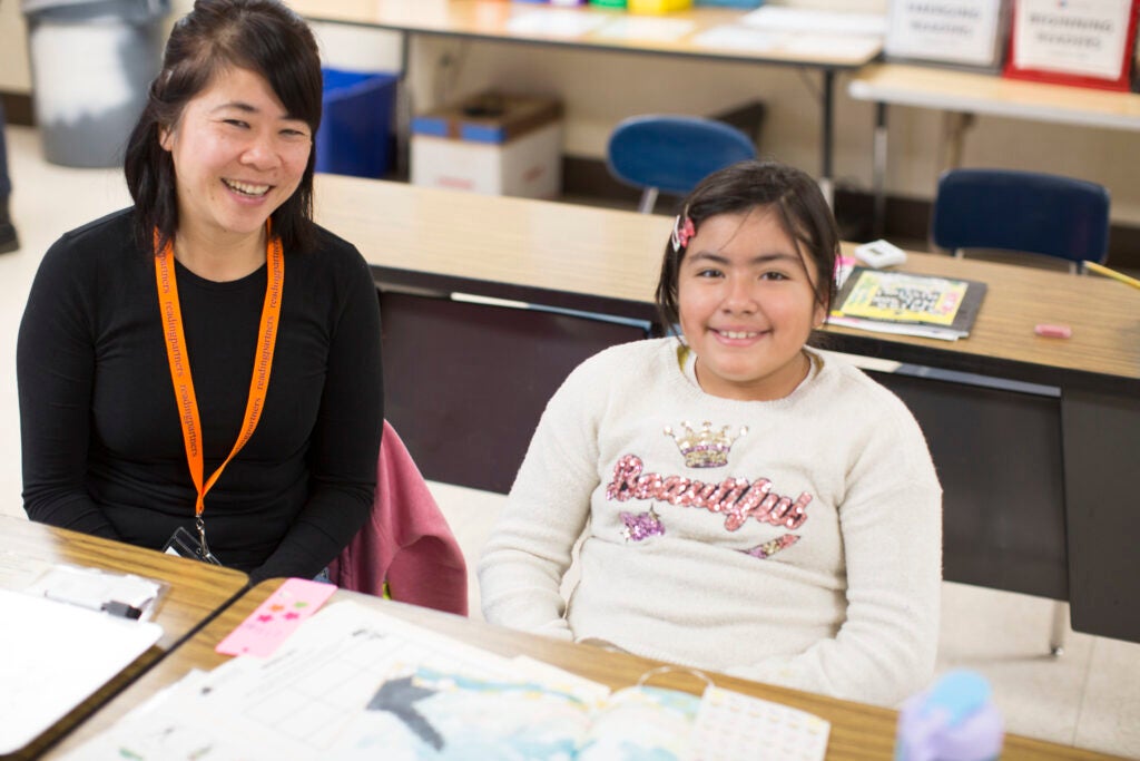 Volunteer tutor and a student sitting at a desk in a classroom side-by-side smiling. 