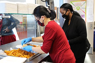 Two cafeteria supervisors prepare meals for students in a school lunch room.