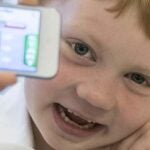 A young student in a classroom holds up a mobile phone and smiles for a photo