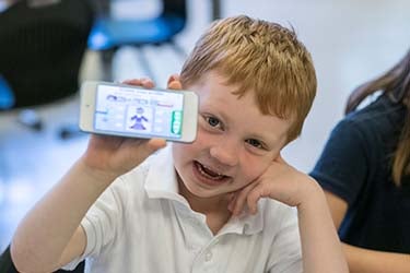 A young student sits in a classroom holding a mobile phone and smiling for a photo.