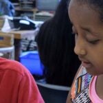 Two students work together at a desk in a classroom.