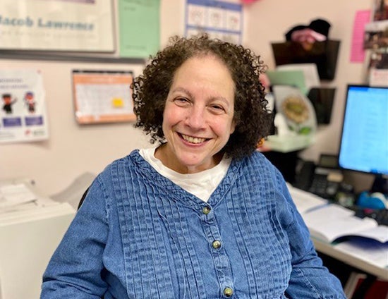 Joan Steinberg smiles for a photo while sitting at a desk in a school office.