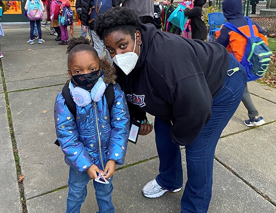 Laticia Bazemore and a young student pose for a photo outside a school while students and adults line up to go inside.