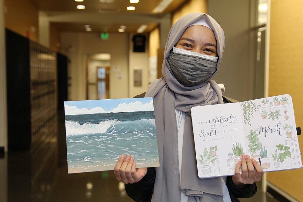 A student stands in a school hallway holding up two pencil drawings.