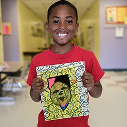 A young student holds up a piece of student art and smiles for a photo