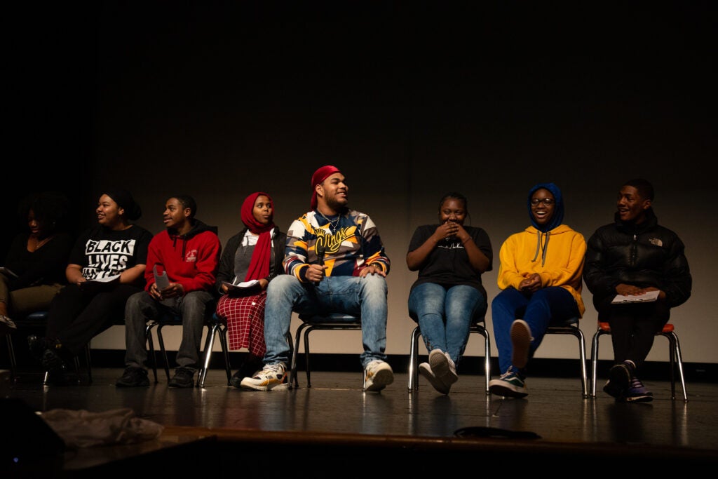 A group of high school students sit on a stage during an event.