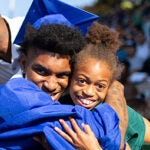 A graduate in cap and grown hugs a younger child at a graduation ceremony.