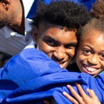 Four people at a graduation ceremony including two graduates in cap and gown hug family members.